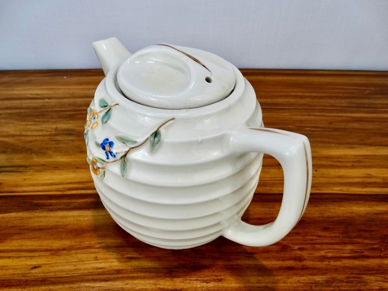 May include: White ceramic teapot with a ribbed design and floral accents. The teapot has a lid with a small hole in the top and a gold-trimmed handle.