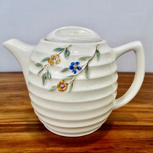 May include: A white ceramic teapot with a ribbed design and floral accents. The teapot has a lid and a handle. The flowers are yellow, blue, and green.