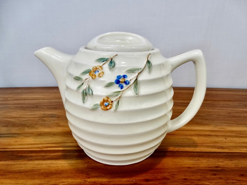 May include: A white ceramic teapot with a ribbed design and floral accents. The teapot has a lid and a handle. The flowers are yellow, blue, and green.