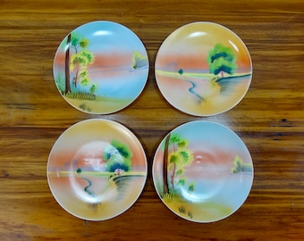 May include: Set of four ceramic plates with a landscape design. Each plate features a different scene with trees, a river, and a sunset sky. The plates are white with a light blue, yellow, and orange glaze.