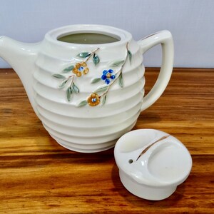 May include: White ceramic teapot with a floral design and a matching lid. The teapot has a ribbed body and a small spout. The lid has a small hole in the centre.
