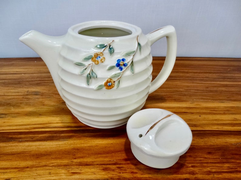 May include: White ceramic teapot with a floral design and a matching lid. The teapot has a ribbed body and a small spout. The lid has a small hole in the centre.