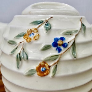 May include: White ceramic teapot with a ribbed design and floral accents. The teapot features a blue and yellow flower design with green leaves.
