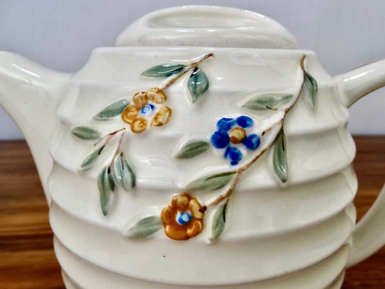 May include: White ceramic teapot with a ribbed design and floral accents. The teapot features a blue and yellow flower design with green leaves.