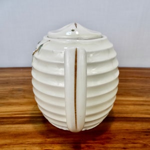May include: White ceramic teapot with a ribbed design and a gold handle. The teapot has a lid with a small hole in the top.