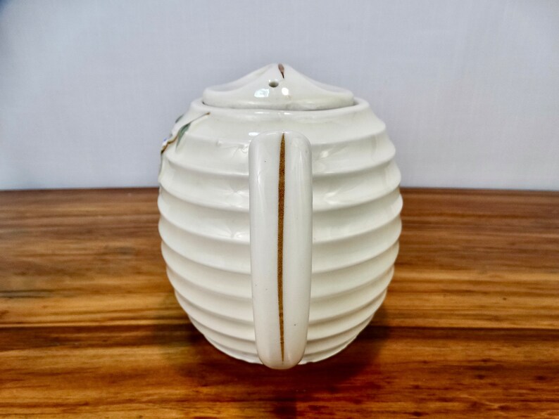 May include: White ceramic teapot with a ribbed design and a gold handle. The teapot has a lid with a small hole in the top.
