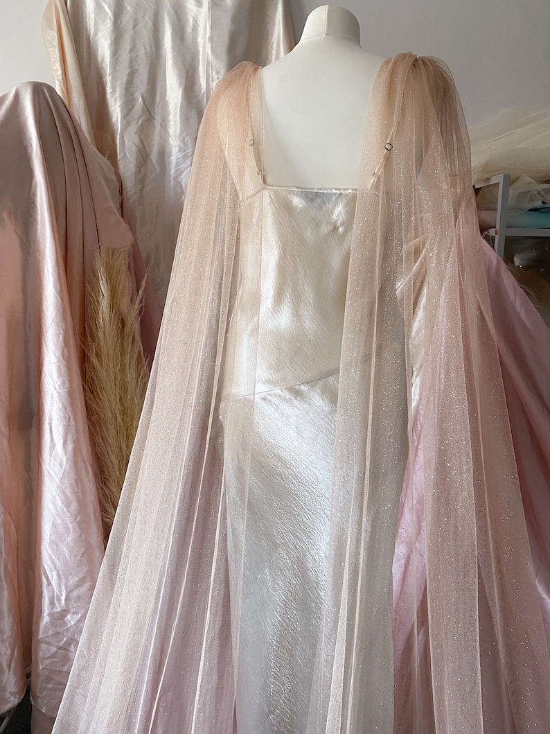 Puede incluir: Un vestido de sat&eacute;n blanco con una superposici&oacute;n de tul rosa y un acabado brillante. El vestido tiene un escote cuadrado y una silueta ajustada.
