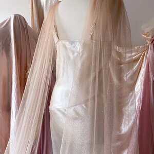 Puede incluir: Un vestido de sat&eacute;n blanco con una superposici&oacute;n de tul rosa brillante. El vestido tiene un escote redondo y una silueta ajustada. La superposici&oacute;n tiene mangas largas y una cola fluida.