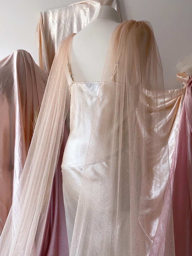 Puede incluir: Un vestido de sat&eacute;n blanco con una superposici&oacute;n de tul rosa brillante. El vestido tiene un escote redondo y una silueta ajustada. La superposici&oacute;n tiene mangas largas y una cola fluida.