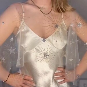 May include: Ivory satin slip dress with a sheer overlay adorned with silver starburst and star accents. The dress features a V-neckline and spaghetti straps. A delicate gold necklace adds a touch of elegance. The overall aesthetic is celestial and glamorous.