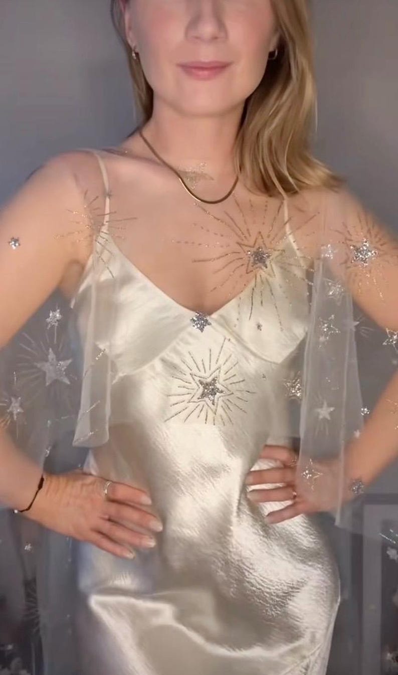 May include: Ivory satin slip dress with a sheer overlay adorned with silver starburst and star accents. The dress features a V-neckline and spaghetti straps. A delicate gold necklace adds a touch of elegance. The overall aesthetic is celestial and glamorous.