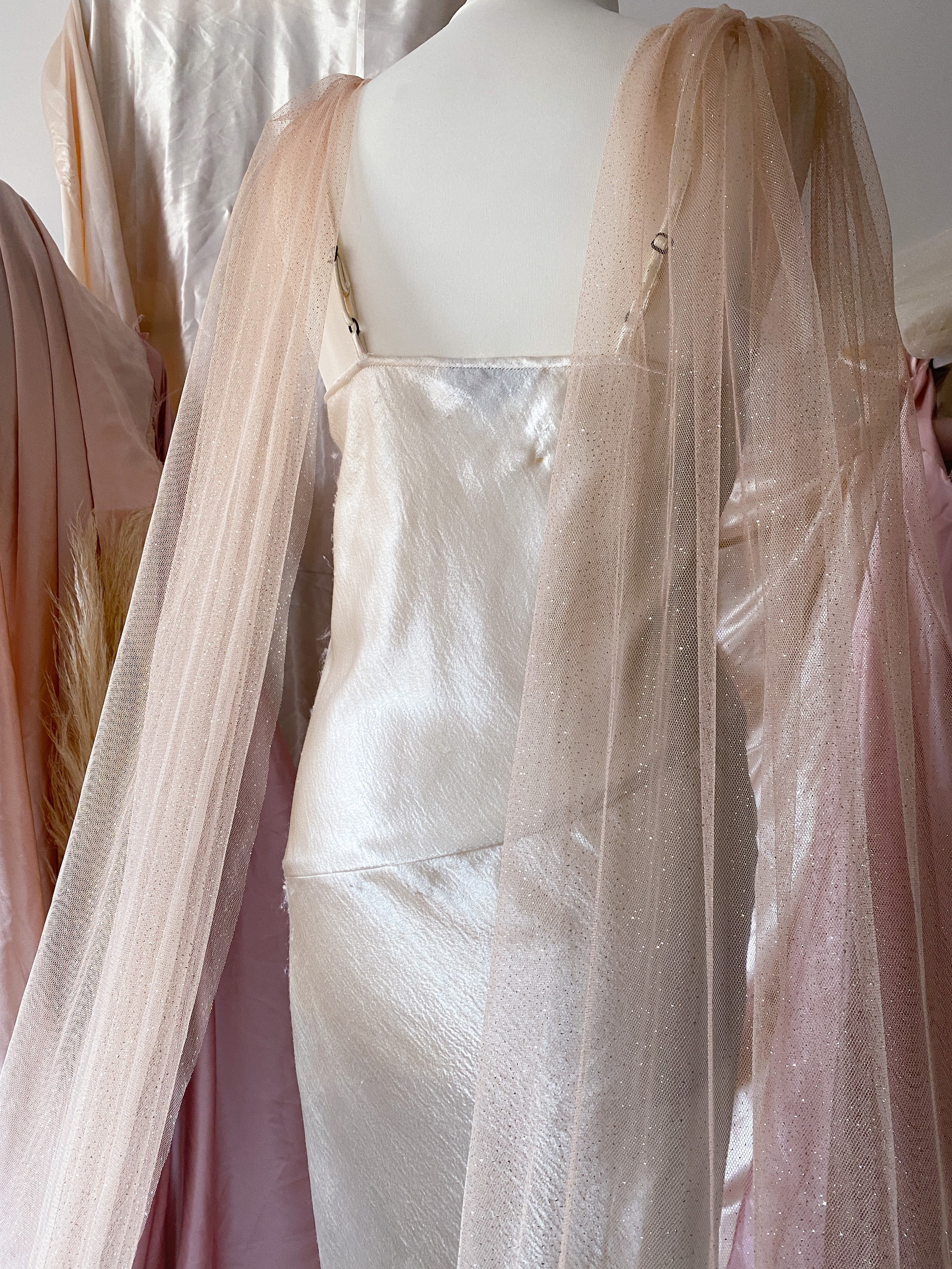 Puede incluir: Un vestido de sat&eacute;n blanco con una superposici&oacute;n transparente y brillante. La superposici&oacute;n tiene un detalle fruncido en los hombros y cae hasta el suelo.
