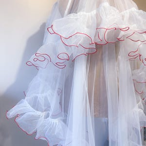 Pode incluir: Uma peça de roupa branca e transparente com babados com acabamento vermelho. Parece ser um roupão ou uma sobreposição, com uma qualidade fluida e etérea. Os babados adicionam um elemento decorativo ao design.