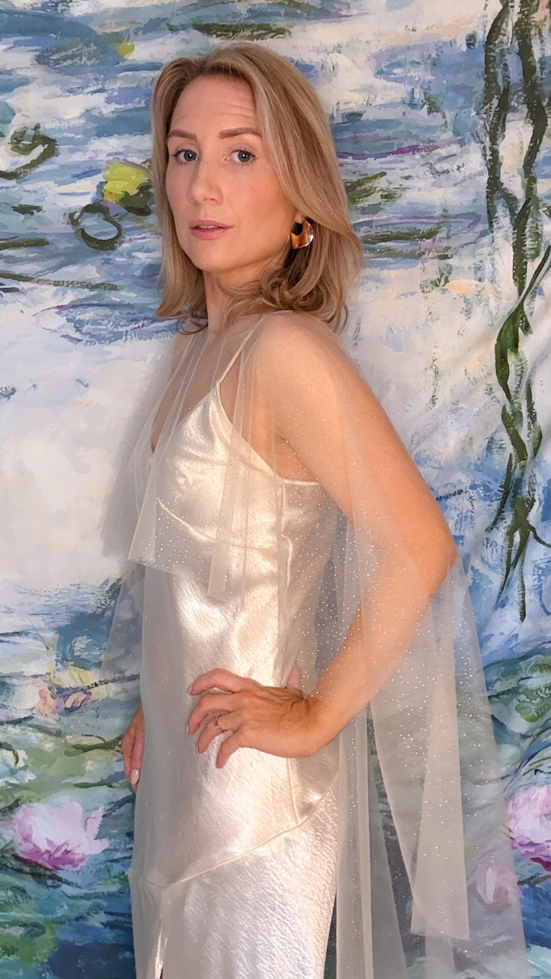 May include: A cream-colored satin slip dress with a sheer, sparkling overlay. The dress has spaghetti straps and a flowing silhouette. The model is wearing large, brown hoop earrings. The background is a colorful abstract painting.