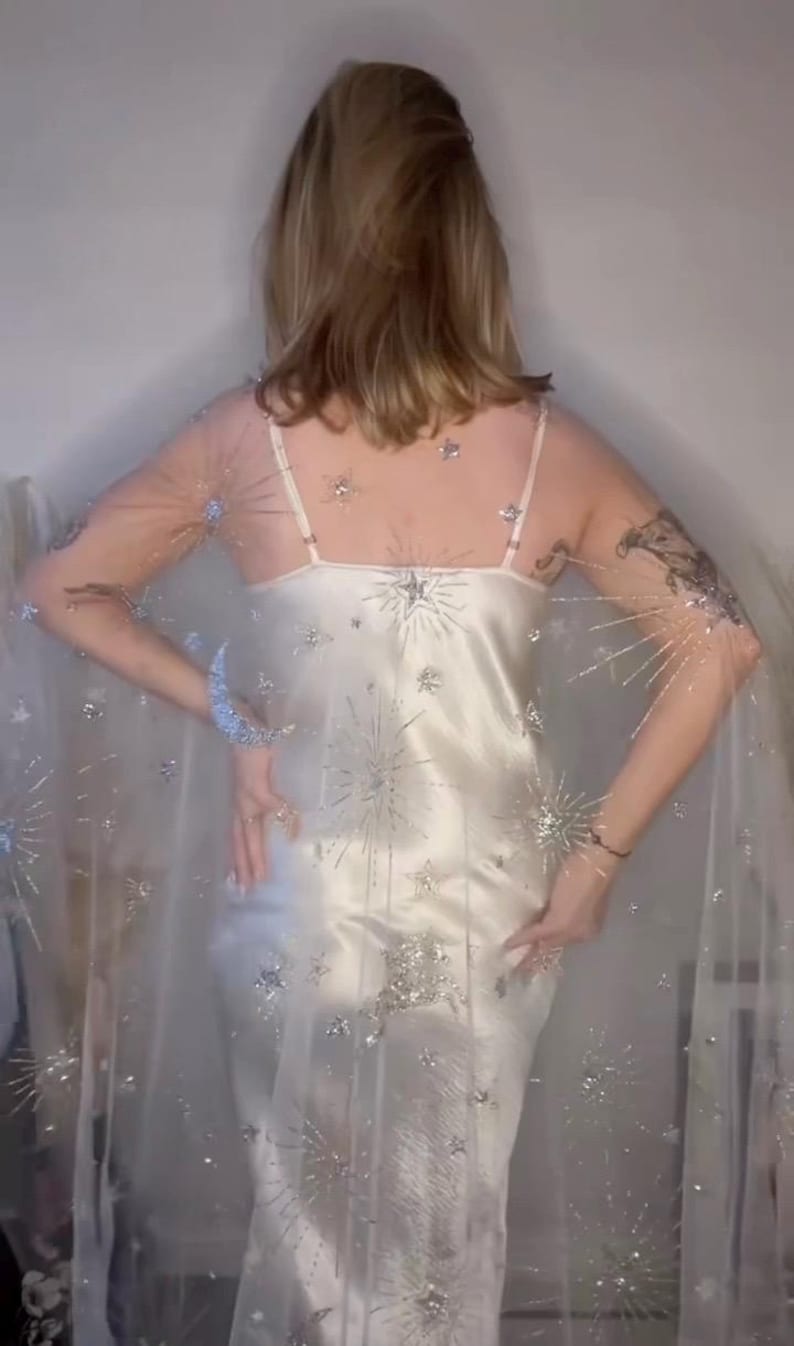 May include: A white satin slip dress with spaghetti straps, overlaid with a sheer cape. The cape and dress are embellished with silver starburst and star-shaped sequins. The dress is floor-length, creating an elegant, celestial aesthetic.