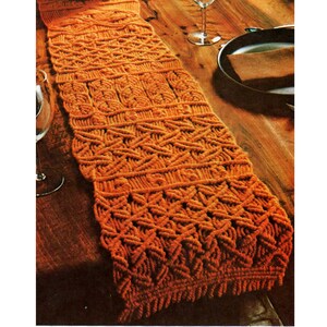 May include: A long, orange macrame table runner with a geometric pattern. The runner is made with thick cord and has a fringe on the ends.