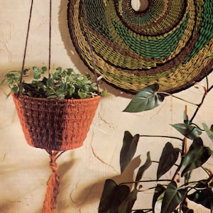 May include: A hanging macrame plant holder with a brown and orange pattern, filled with green plants. A large round macrame wall hanging with a green, brown, and black pattern hangs on the wall behind the plant holder.