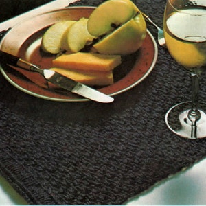 May include: A black crocheted placemat with a brown plate, a knife, and a glass of white wine. The plate contains sliced apples and cheese.