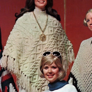 Puede incluir: Dos mujeres con ponchos de punto blanco con flecos. Una mujer lleva un collar de oro y la otra lleva gafas de sol. Las mujeres están de pie frente a un fondo rojo.