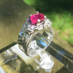 May include: A silver ring with a large, oval, bright pink gemstone. The ring has an ornate, detailed design around the gemstone. The ring is displayed on a clear plastic stand, with a blurred green background.