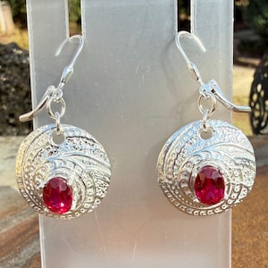 May include: A pair of silver-toned earrings. Each earring features a circular, textured design with an oval, ruby-red gemstone at the center. The earrings have a lever-back closure.