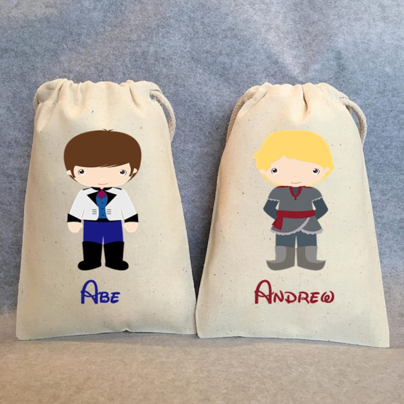 Puede incluir: Dos bolsas de tela blanca con cord&oacute;n de ajuste y nombres personalizados. Una bolsa presenta un ni&ntilde;o de dibujos animados con cabello casta&ntilde;o que lleva un atuendo azul y blanco. La otra bolsa presenta un ni&ntilde;o de dibujos animados con cabello rubio que lleva un atuendo gris y negro. Los nombres "Abe" y "Andrew" est&aacute;n bordados en las bolsas.