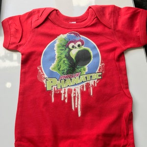 Peut inclure: Combinaison bébé rouge avec un graphique vert et bleu du mascotte Phillie Phanatic. Le graphique a le texte "PHILLIE PHANATIC" en blanc.