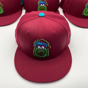 Könnte beinhalten: Eine marineblaue Baseballkappe mit einem grünen, blauen und violetten Monstercharakter, der auf der Vorderseite gestickt ist. Das Monster trägt ein blaues und rotes Bandana auf dem Kopf.