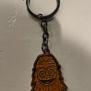 Op de afbeelding: Een bruine metalen sleutelhanger met een cartoonfiguur met een brede glimlach, grote ogen en een borstelige baard. De sleutelhanger is bevestigd aan een zilveren metalen ring en ketting.
