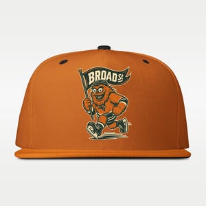 Op de afbeelding: Oranje baseballpet met platte klep met een cartoonmascotte die rent met een vlag met de tekst "BROAD St.". De mascotte is oranje met zwarte en witte accenten, met schaatsen. De pet heeft een zwarte knop aan de bovenkant.
