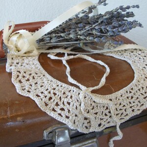 May include: A cream-colored, crocheted collar with a scalloped edge rests on a vintage brown suitcase. A bundle of dried lavender and a cream ribbon with a fabric flower are attached to the suitcase handle.