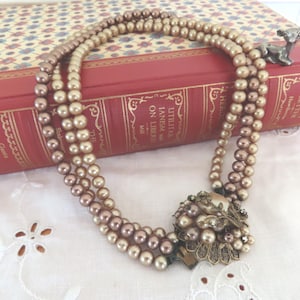 May include: A multi-strand necklace featuring lustrous champagne and bronze-toned pearls. The necklace is secured with a decorative, ornate metal clasp adorned with smaller pearls and clear crystals, creating a focal point on the strand.