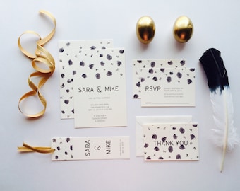 May include: A wedding stationery set with a cream background and dark grey watercolour splotches. Includes invitations, RSVP cards, thank you cards, and a bookmark. Gold ribbon, two gold eggs, and a black-tipped feather complete the arrangement.