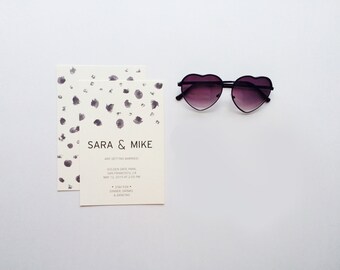 May include: Wedding invitations with the names "SARA & MIKE" and event details, next to heart-shaped sunglasses with gradient lenses. The invitations have a cream background with dark purple watercolour splotches.