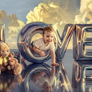 May include: A toddler in a white t-shirt crawls through the center of a large, metallic, silver-colored letter O that is part of a larger LOVE sign. Next to the sign sits a large brown teddy bear holding a smaller brown teddy bear with a yellow ribbon.