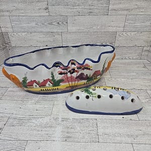 May include: An oval ceramic bowl with a scalloped rim and a matching removable lid featuring multiple circular holes. The exterior is decorated with a hand-painted landscape scene of houses, trees, and mountains in red, green, and yellow tones.