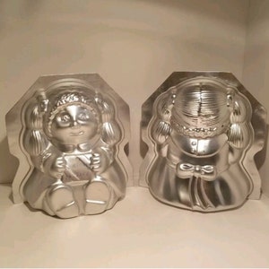 Puede incluir: Dos moldes para pasteles de metal plateado con forma de muñecas. Las muñecas llevan vestidos y tienen coletas. La muñeca de la izquierda sostiene un libro que dice "Birthday Wishes".