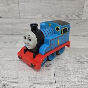 May include: A blue, red, and black toy train with the number 1 on the side. The train has a smiling face and is made of plastic. The toy train is a children's toy.
