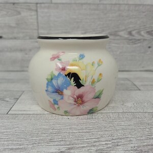 Puede incluir: Un tarro de cerámica blanco con un borde negro y un diseño floral con flores rosas, azules, amarillas y verdes.