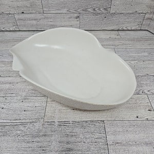 Puede incluir: Una fuente blanca en forma de riñón con un borde texturizado. La fuente tiene un interior liso y brillante y un pequeño pico en un lado. La fuente está hecha de cerámica o porcelana y es adecuada para servir alimentos.