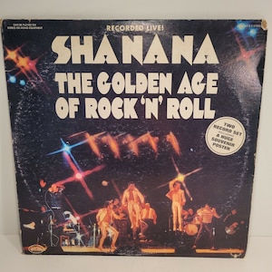 May include: Vintage vinyl record album cover for "Shanana: The Golden Age of Rock 'N' Roll." The cover is dark blue with the band's name and album title in large white letters. The image includes a live performance scene with musicians and stage lights.