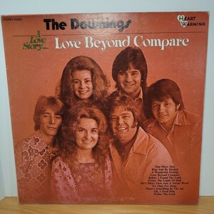 May include: A vintage vinyl record album cover featuring a group of six people, four men and two women, with the title "The Downings: A Love Story... Love Beyond Compare" and the words "Heart Warming" in a heart shape. The album cover is orange and features a photo of the group in a studio setting.