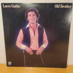 May include: Album cover for "Oh! Brother" by Larry Gatlin. The cover features a smiling man in a pink shirt, black vest, and black pants. The album title and artist's name are printed in white on a black background.