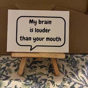 May include: A rectangular white card featuring a black speech bubble outline and the black text "My brain is louder than your mouth" displayed on a small wooden easel. The card has a textured surface and is positioned in a horizontal orientation.