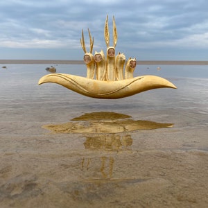 May include: A golden sculpture of a boat with five figures with animal heads, floating above the water on a sandy beach.