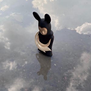 May include: A black ceramic sculpture of a figure with large ears and a white ceramic piece wrapped around its body. The sculpture is standing in a puddle of water, reflecting the sky.