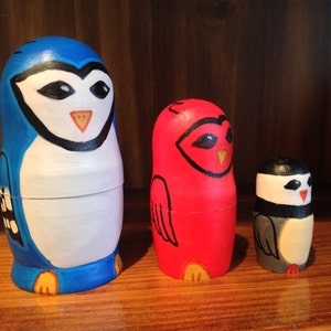 Può includere: Tre bambole russe in legno dipinte a mano, o bambole matrioska, che raffigurano un pinguino blu e bianco, un cardinale rosso e un pinguino più piccolo in bianco e nero.