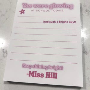 May include: A white rectangular notepad with pink text and lines. The header reads You were glowing AT SCHOOL TODAY! with a small flower icon. The text had such a bright day!! is written on the first line. The footer says Keep shining bright! -Miss Hill.