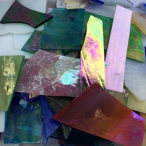 May include: A collection of assorted stained glass pieces in various shapes and sizes. The fragments feature a diverse range of colors including iridescent pink, deep blue, emerald green, and opaque white, with some pieces displaying a metallic finish.