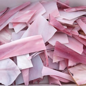 May include: A collection of irregularly shaped, pink stained glass pieces. The glass shards vary in shades of pink, from pale to deep rose, and are arranged in a container. The pieces have a translucent quality.